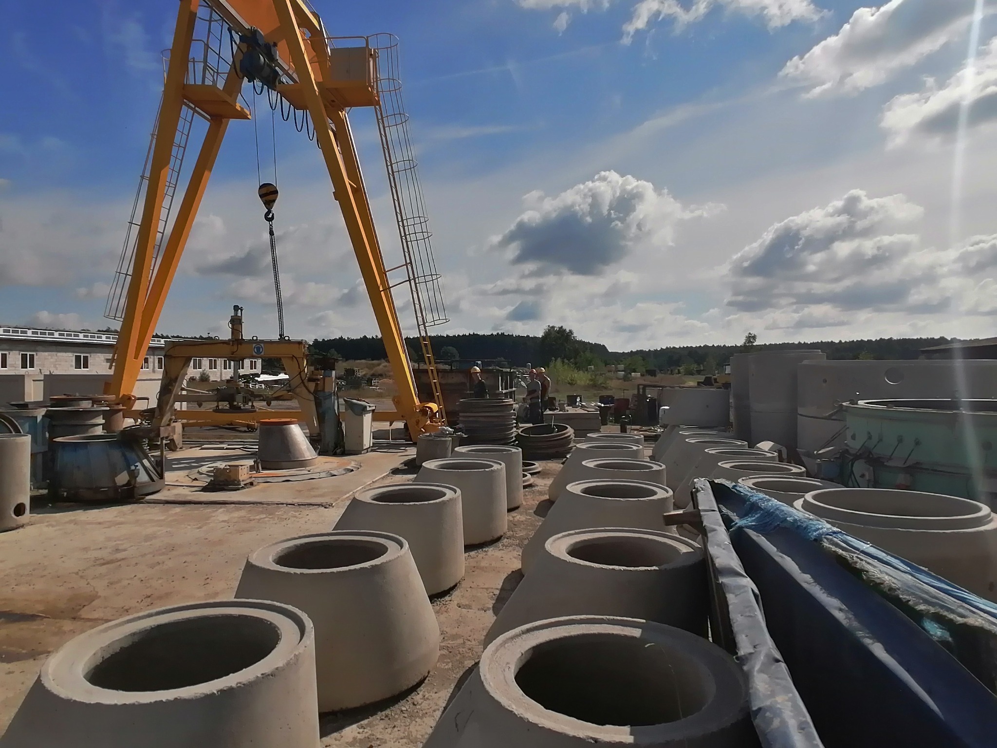 Betonowe kręgi ustawione w rzędach na placu budowy, żółty dźwig bramowy w tle, pracownicy w kaskach w oddali, błękitne niebo z chmurami.