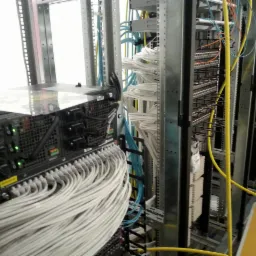 Serwerownia z zainstalowanym sprzętem, widoczne liczne kable sieciowe w kolorze białym, żółtym i niebieskim, podłączone do paneli krosowych i urządzeń sieciowych w metalowej szafie rack.