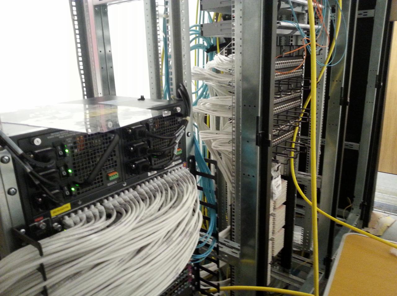 Serwerownia z zainstalowanym sprzętem, widoczne liczne kable sieciowe w kolorze białym, żółtym i niebieskim, podłączone do paneli krosowych i urządzeń sieciowych w metalowej szafie rack.
