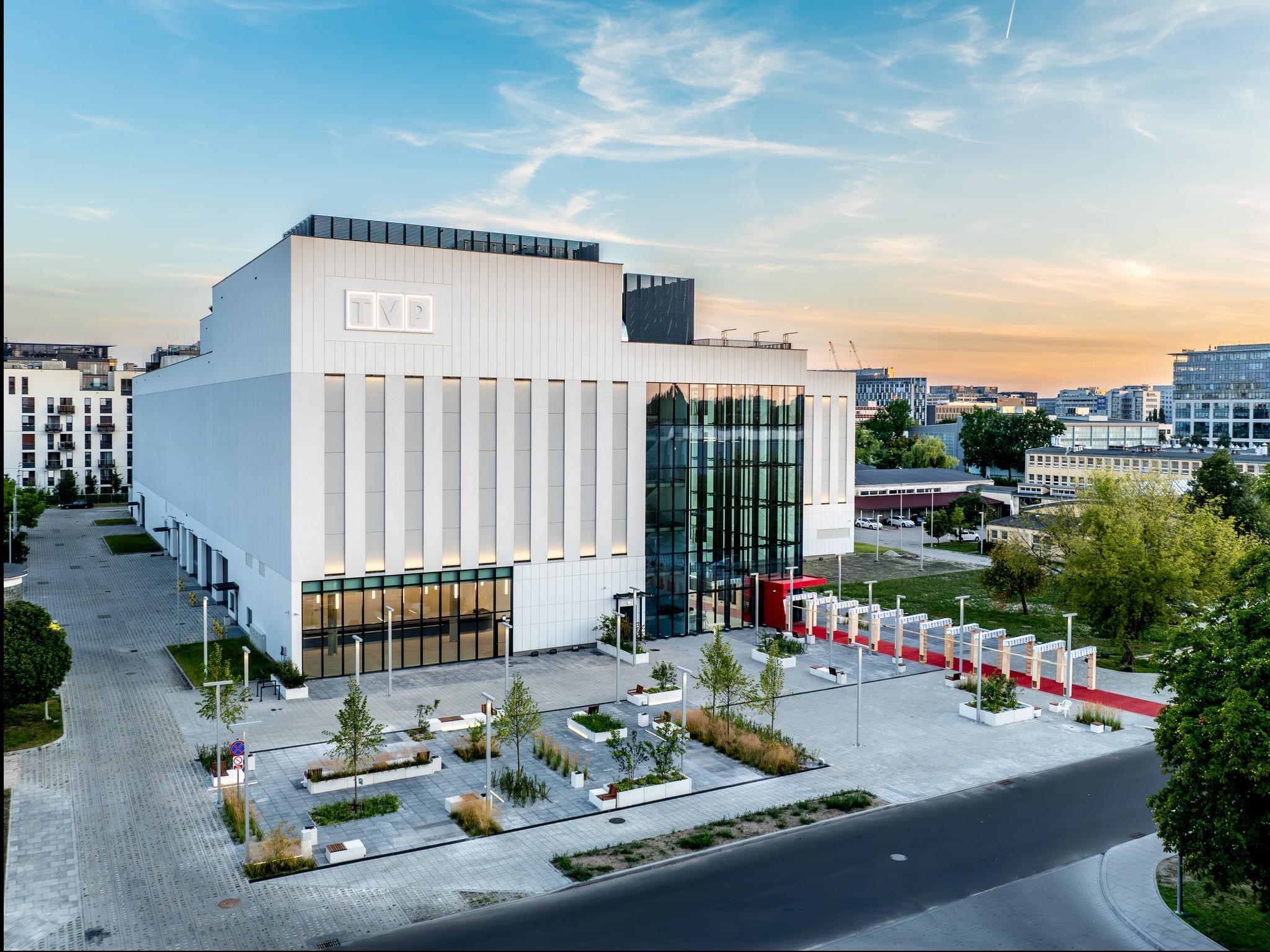 Nowoczesny budynek TVP w minimalistycznym stylu, z elementami szkła i betonu, otoczony zielenią i przestrzenią publiczną. Ujęcie z lotu ptaka, wieczorne oświetlenie.