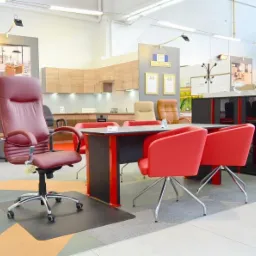 Ekspozycja krzeseł biurowych: bordowy fotel obrotowy na czarnej macie oraz czerwone krzesła konferencyjne przy stole w salonie meblowym z ekspozycją kuchni i szaf w tle.