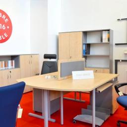 Biuro i Meble B.M. Marek Józef Mikołowski - Jasne biuro z biurkiem o zaokrąglonym blacie, krzesłami w kolorze niebieskim i czarnym, oraz szafami i regałami z jasnego drewna. Na ścianie widoczny okrągły znak 'Dobre Meble 2016'. Na biurku...