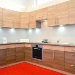 Biuro i Meble B.M. Marek Józef Mikołowski - Kątowe ustawienie jasnych szafek kuchennych z drewnianymi frontami, oświetleniem podszafkowym i czerwonym dywanem na podłodze.