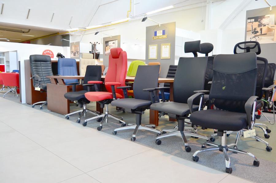 Ekspozycja różnorodnych krzeseł biurowych na kółkach w salonie meblowym, widoczne modele z wysokim oparciem, regulowanymi podłokietnikami i zagłówkami, ustawione rzędem na jasnej podłodze.