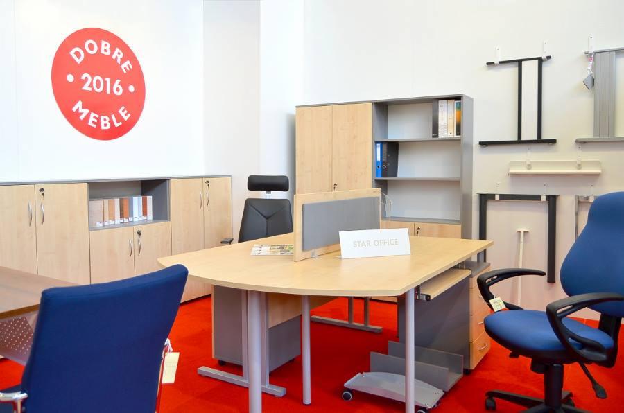 Jasne biuro z biurkiem o zaokrąglonym blacie, krzesłami w kolorze niebieskim i czarnym, oraz szafami i regałami z jasnego drewna. Na ścianie widoczny okrągły znak 'Dobre Meble 2016'. Na biurku...