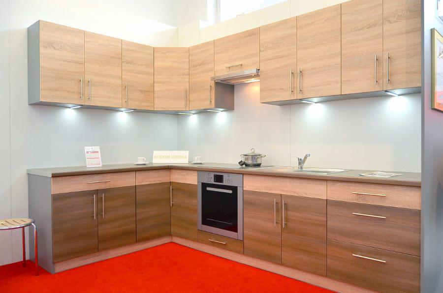 Kątowe ustawienie jasnych szafek kuchennych z drewnianymi frontami, oświetleniem podszafkowym i czerwonym dywanem na podłodze.