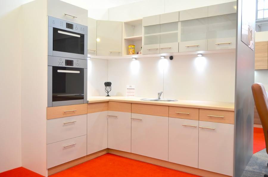 Nowoczesna kuchnia z białymi szafkami dolnymi i górnymi z przeszklonymi frontami, drewnianym blatem, dwoma piekarnikami w zabudowie i jasnym oświetleniem punktowym, na podłodze pomarańczowa...