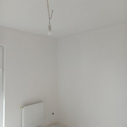 Kraw-bud - Pusty, jasny pokój po remoncie z białymi ścianami, grzejnikiem i gołą żarówką zwisającą z sufitu, widoczne ślady po szpachlowaniu na podłodze.