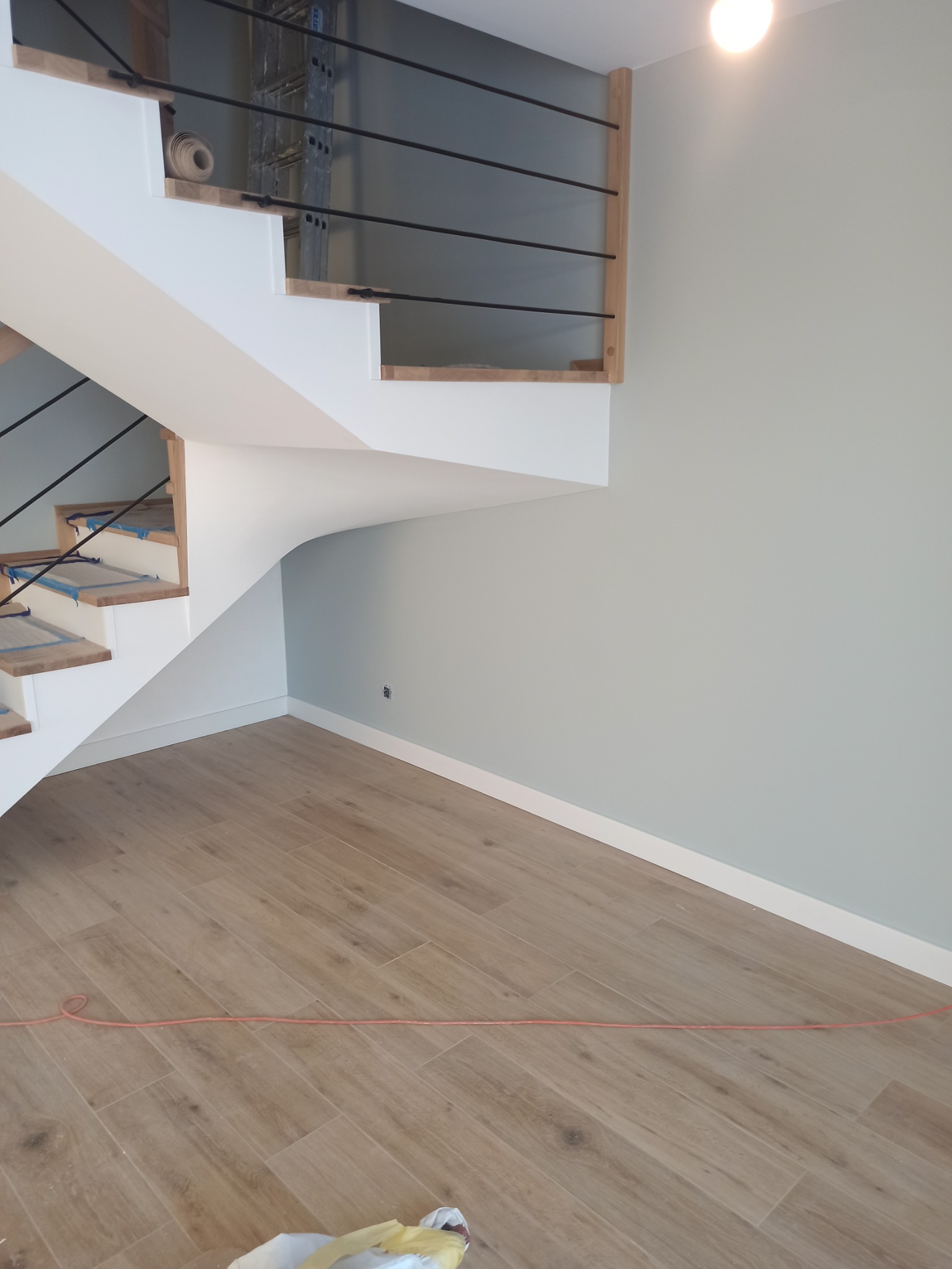 Świeżo pomalowane, jasnoszare ściany w pomieszczeniu ze schodami z drewnianymi stopniami zabezpieczonymi folią, widoczny kabel na podłodze z płytek imitujących drewno.