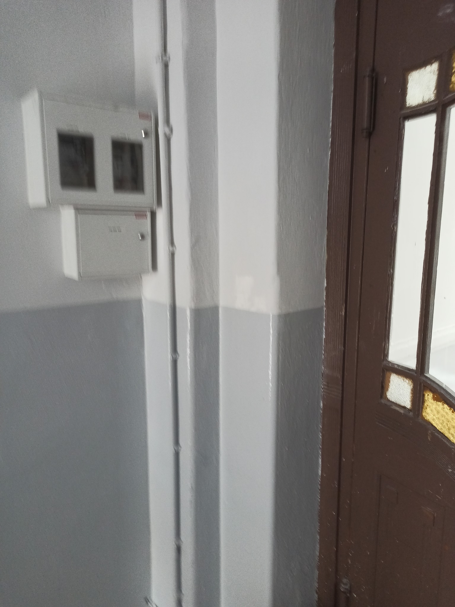 Klatka schodowa z widoczną szarą dolną częścią ściany, białą górną częścią, brązowymi drzwiami z szybkami i skrzynką elektryczną na ścianie.