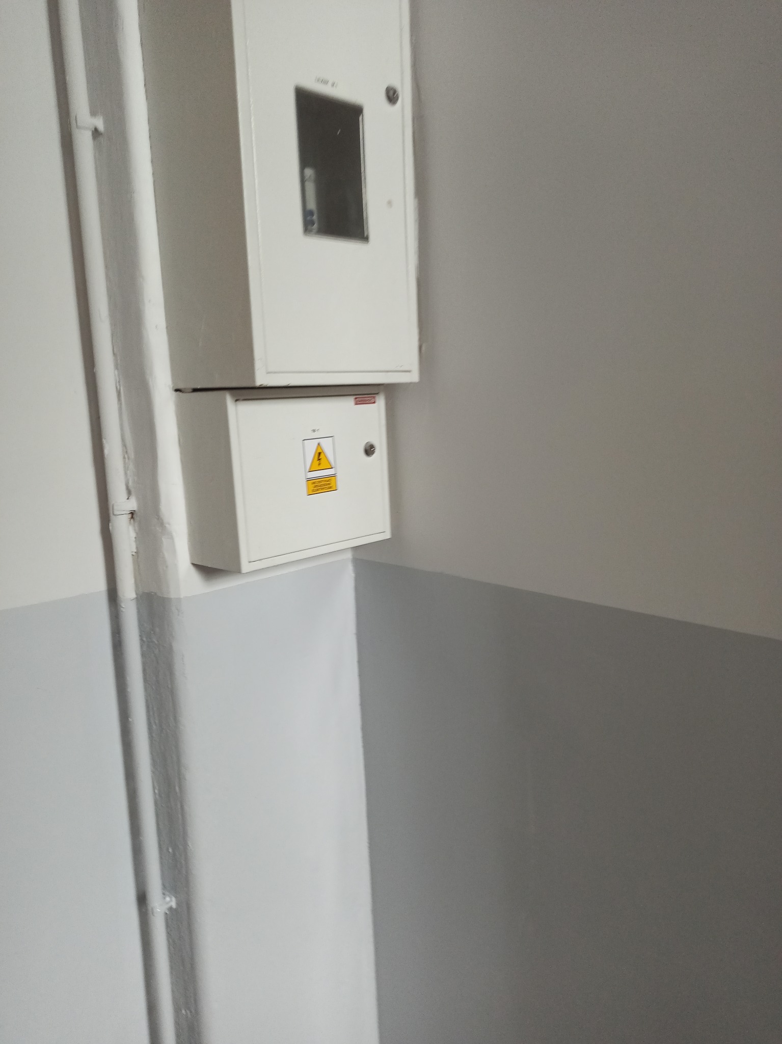 Dwa białe panele elektryczne zamontowane na ścianie, dolna część ściany pomalowana na szaro, widoczne rury instalacyjne wzdłuż ściany.