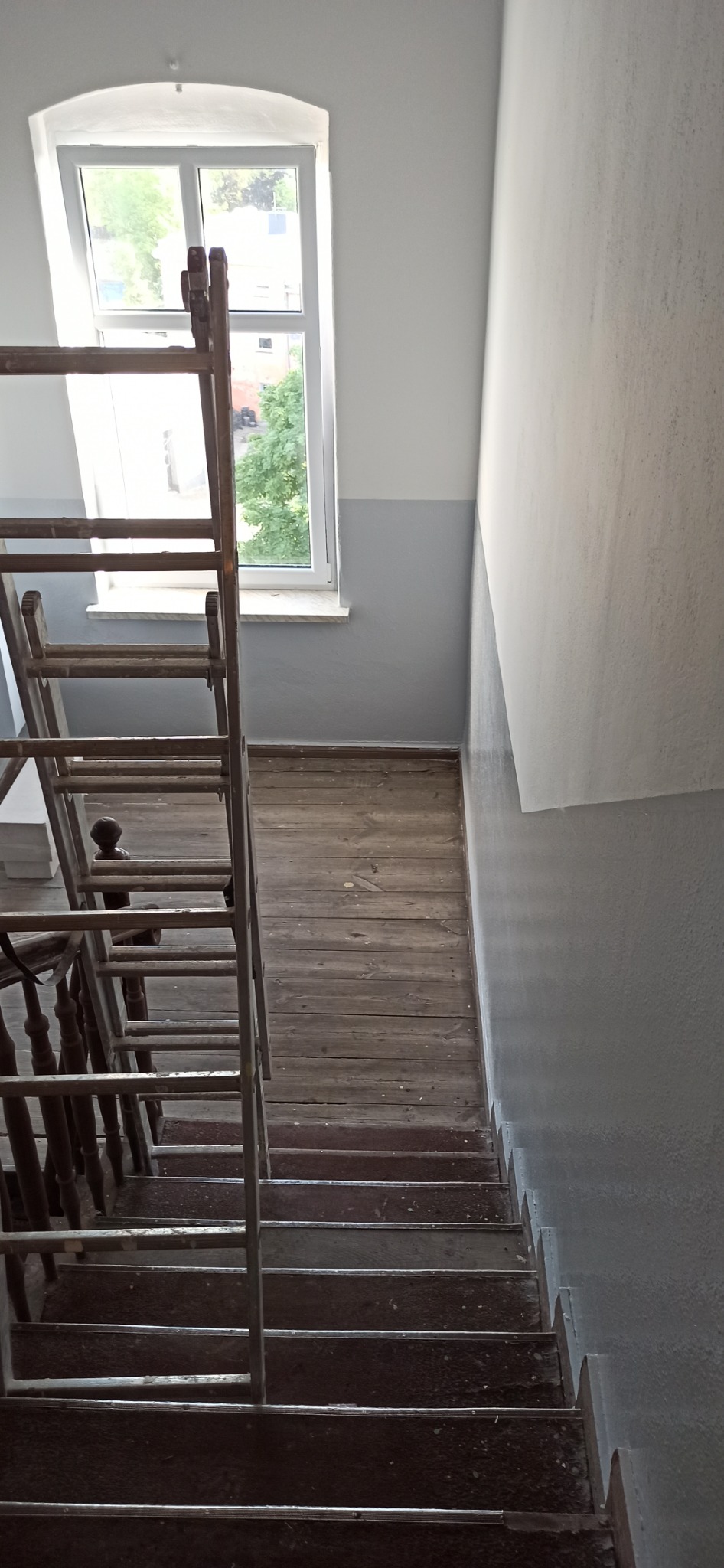 Klatka schodowa z drewnianymi schodami i drabina malarska, ściany pomalowane do połowy na szaro, widok z góry.