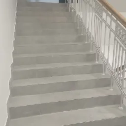 Wykończone schody wewnętrzne z szarych płytek, z białą metalową balustradą i drewnianym poręczą, widok z dołu.