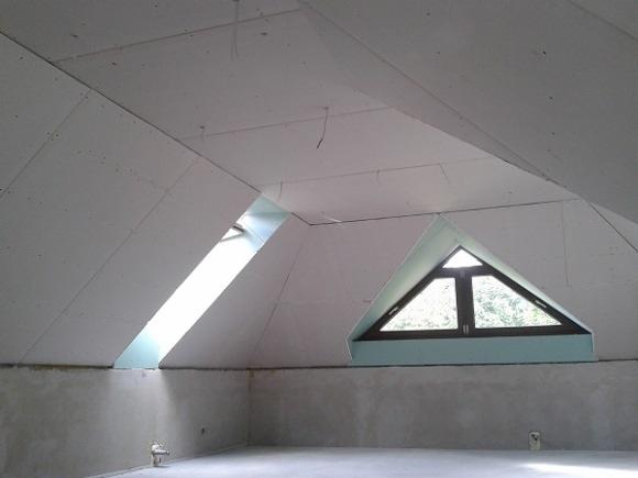 Poddasze w trakcie wykańczania, ściany i sufit pokryte szarą płytą, widoczne okno dachowe trójkątne i prostokątne w skosie dachu, surowy beton na dole ścian.