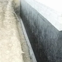 Wykonany wykop wzdłuż fundamentu budynku, zabezpieczonego czarną izolacją przeciwwodną, przygotowany do drenażu.