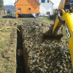 Żółta koparka JCB podczas wykonywania głębokiego wykopu liniowego w ziemi, widoczny żółty przewód w wykopie oraz zabudowa jednorodzinna w tle.