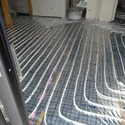 Rozłożone rury ogrzewania podłogowego na folii z nadrukowaną siatką, widoczne elementy konstrukcyjne budynku w tle.