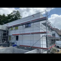 Nowoczesny dom w Grzegorzewie z białą elewacją, otoczony rusztowaniami, widoczne okna z żółtymi foliami ochronnymi oraz niezabudowany jeszcze teren wokół.