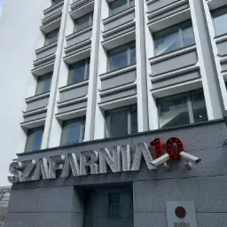 Fasada budynku z nazwą 'Szafarnia 10' wykonaną z liter z żarówkami, uzupełniona o dwie kamery monitoringu.