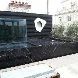 Czarny marmurowy zbiornik wodny z szklaną ścianą na tarasie, z dekoracyjnym panelem z białym geometrycznym logo i roślinami w tle.