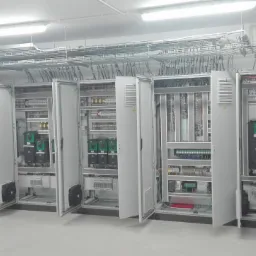 Rozbudowana szafa sterownicza z otwartymi drzwiami, widoczne liczne kable, sterowniki i zabezpieczenia, instalacja kablowa pod sufitem, gaśnica w tle.