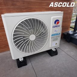ASCOLD - Zewnętrzna jednostka klimatyzacji Gree, zamontowana na szarym tarasie z drewnianym tłem. Widoczny logotyp Ascold i dane kontaktowe firmy.