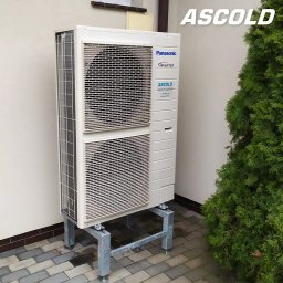 ASCOLD - Zewnętrzna jednostka pompy ciepła Panasonic Inverter na metalowej konstrukcji, ustawiona przy ścianie budynku obok zielonego krzewu. Widoczny bruk.