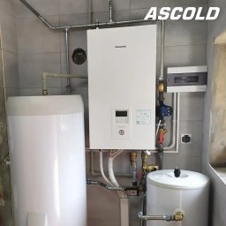 ASCOLD - Biała pompa ciepła Panasonic Aquarea Smart Cloud z dwoma zbiornikami w kotłowni, widoczne rury i instalacja elektryczna na tle szarych płytek.