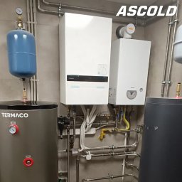 ASCOLD - Dwa białe kotły grzewcze Panasonic i Termet, zbiorniki Termaco i Tigomp, instalacja rurowa na tle szarej ściany. Widoczne logo Ascold w prawym górnym rogu.