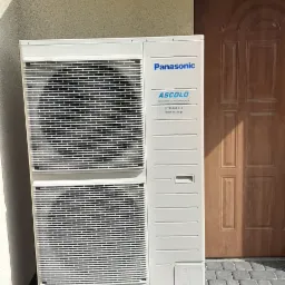 Zewnętrzna jednostka klimatyzacji Panasonic zamontowana na czerwonych betonowych wspornikach przed beżową ścianą domu z drewnianymi drzwiami i kamienną kostką brukową.