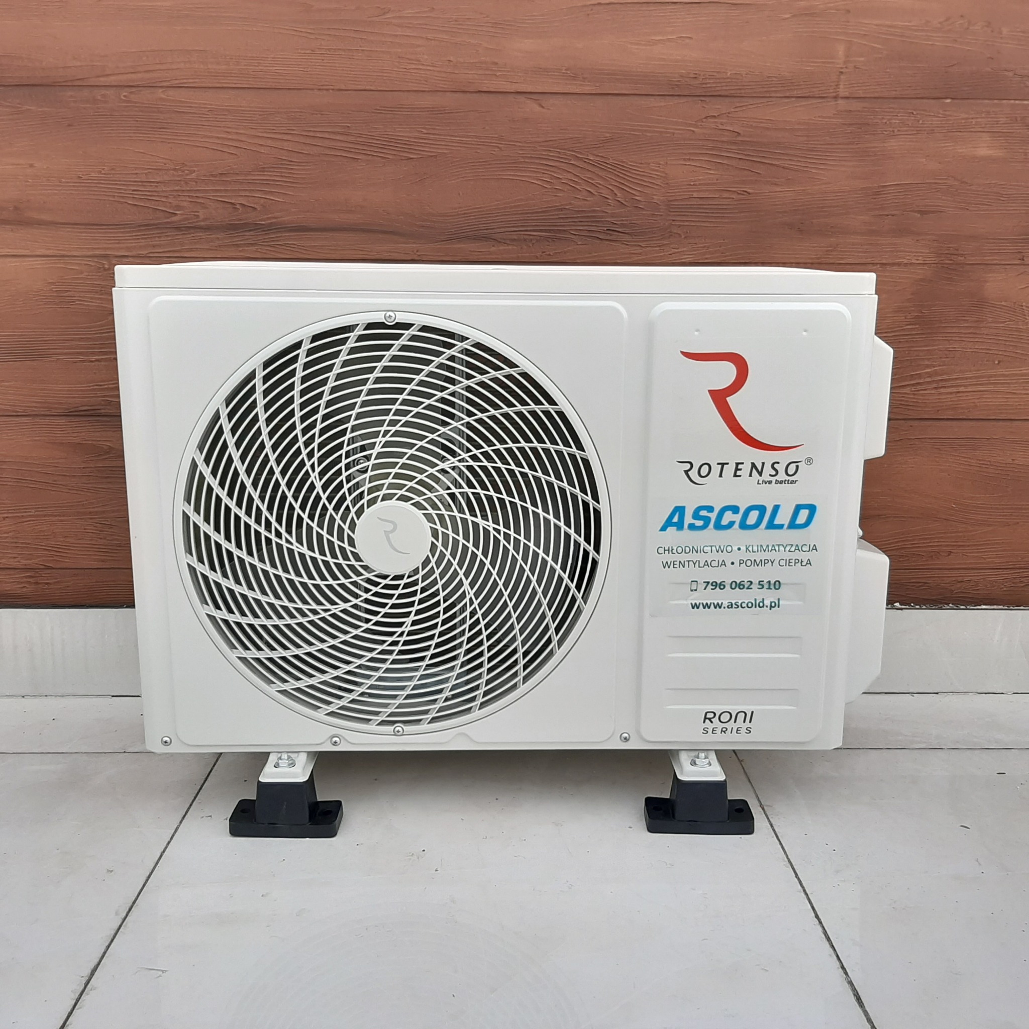 Zewnętrzna jednostka klimatyzacji Rotenso Ascold ustawiona na białych płytkach, z widocznym logo i danymi kontaktowymi firmy Ascold, na tle brązowej ściany imitującej drewno.