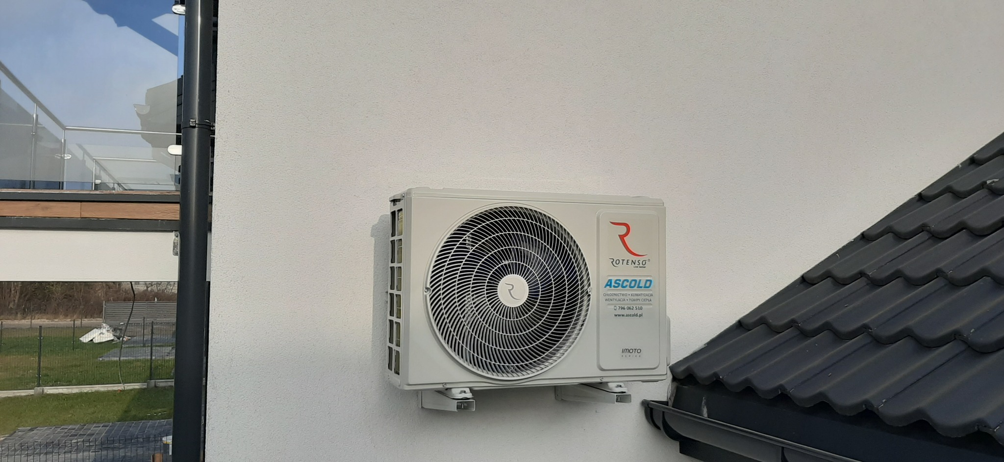 Zewnętrzna jednostka klimatyzacji Rotenso Imoto zamontowana na elewacji budynku z widocznym fragmentem dachu i rynny.