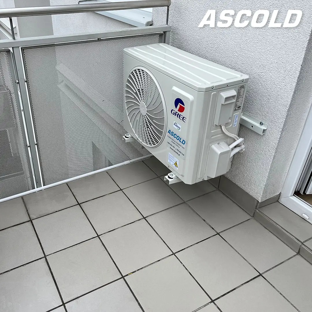 Biała jednostka zewnętrzna klimatyzacji Gree zamontowana na balkonie z szarą balustradą i płytkami podłogowymi. Widoczne mocowania i fragment ściany budynku.