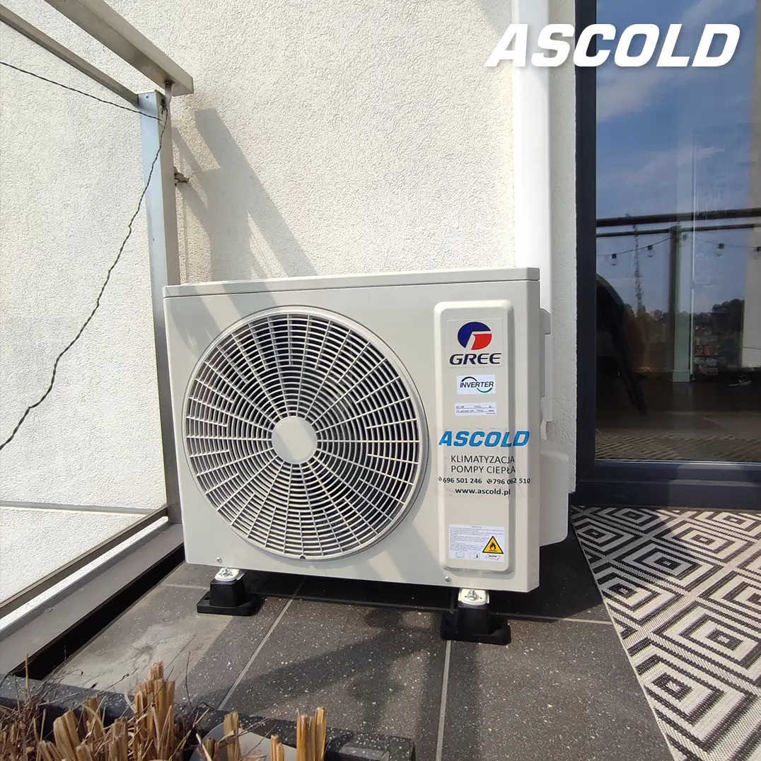 Biała jednostka zewnętrzna klimatyzacji Gree, zamontowana na balkonie z czarną podłogą i wzorzystym dywanikiem. Widoczny fragment elewacji i okna balkonowego.