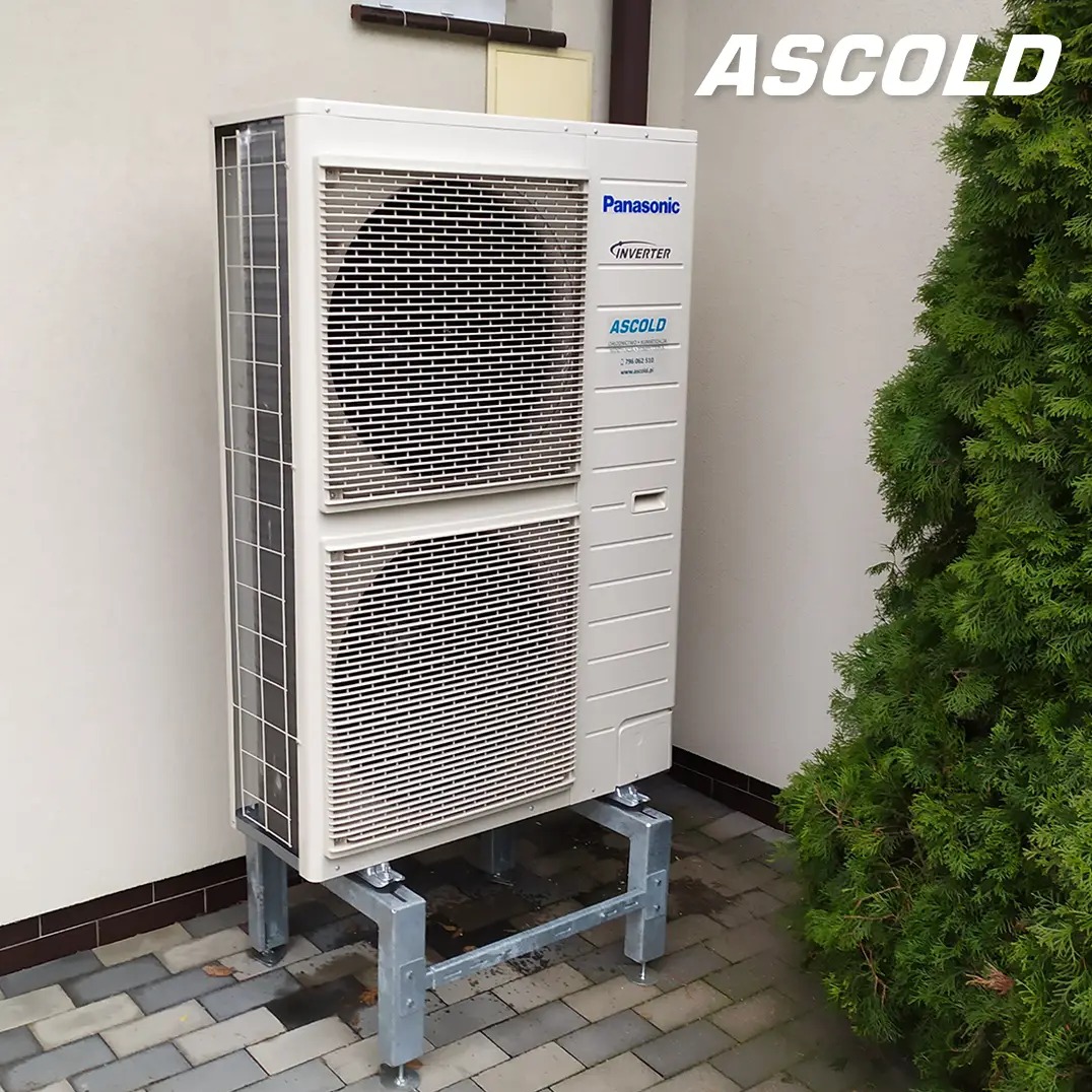 Zewnętrzna jednostka pompy ciepła Panasonic Inverter na metalowej konstrukcji, ustawiona przy ścianie budynku obok zielonego krzewu. Widoczny bruk.