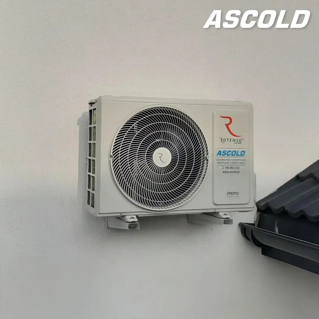 Biała jednostka zewnętrzna klimatyzacji Rotenso Ascold zamontowana na ścianie budynku, widoczny fragment dachu. Minimalistyczny design, nowoczesne rozwiązanie.