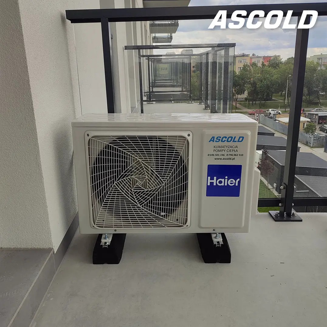 Biała jednostka zewnętrzna klimatyzacji Haier na balkonie z szklaną balustradą. Widoczne logo Ascold i dane kontaktowe. Tło stanowią budynki mieszkalne i zieleń.
