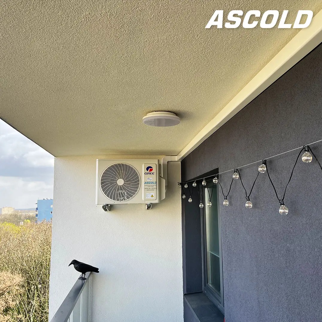 Biała klimatyzacja Gree zamontowana na balkonie z widokiem na miasto, oświetlona girlandą. Na balustradzie siedzi czarny ptak. Widoczne logo Ascold.