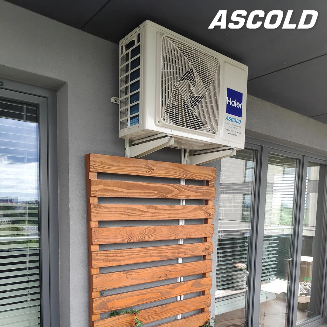 Biała klimatyzacja Haier zamontowana na szarej ścianie balkonu, z drewnianą osłoną z palet. Widok z dołu, okno i przesuwne drzwi balkonowe w tle.