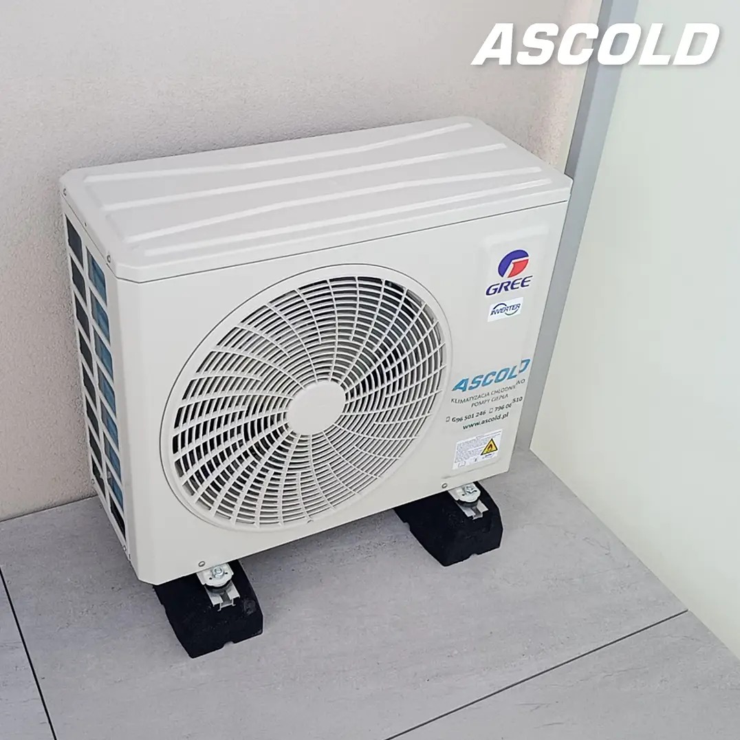 Biały klimatyzator GREE Inverter na czarnych podkładkach antywibracyjnych, ustawiony w rogu pomieszczenia z szarą podłogą i jasnymi ścianami. Widoczne logo Ascold.