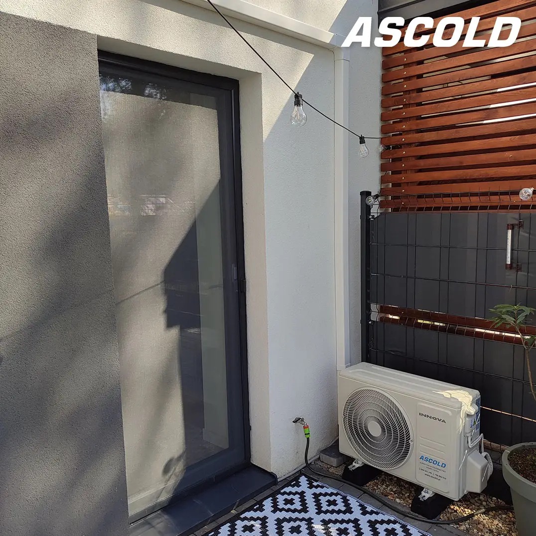 Biała klimatyzacja Innova zamontowana na zewnątrz budynku, obok okna i dekoracyjnej drewnianej ścianki. Widoczne oświetlenie sznurkowe i wzorzysty dywanik.