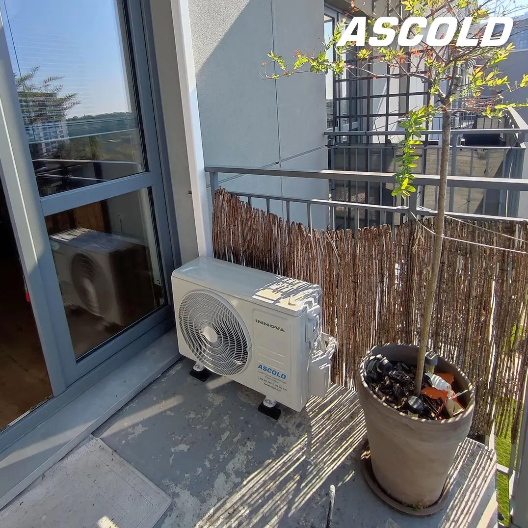 Biała klimatyzacja Innova Ascold na balkonie z szarą podłogą i bambusową osłoną, obok donica z drzewkiem. Widok na miasto w tle.