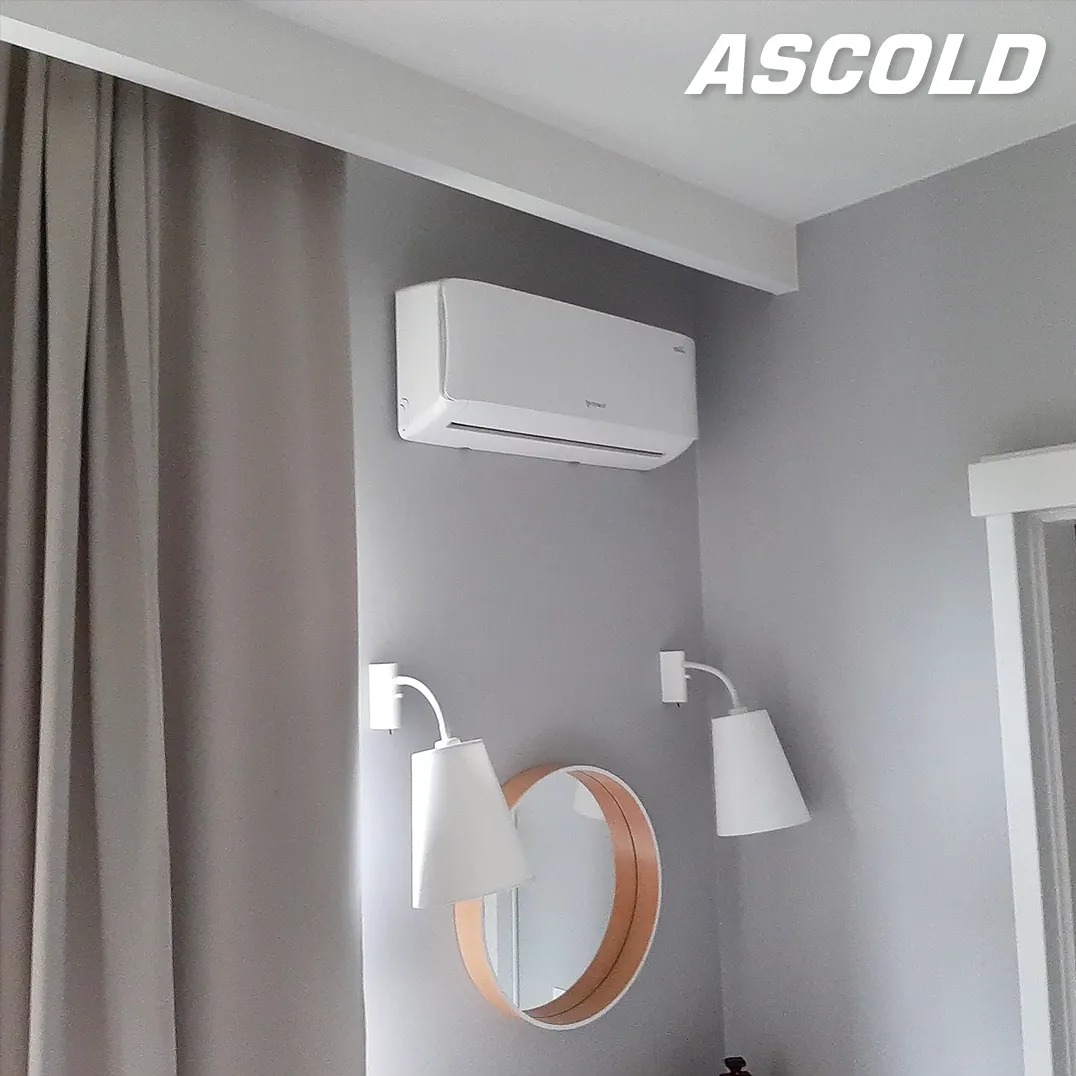 Biały klimatyzator Ascold na szarej ścianie w pokoju, z lampkami i okrągłym lustrem. Widoczne zasłony i fragment drzwi. Minimalistyczny design.
