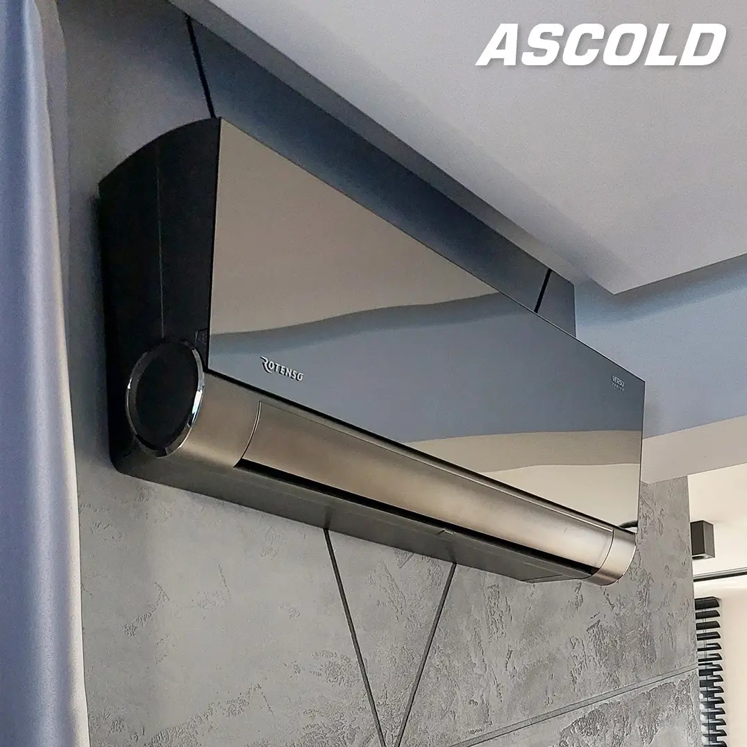 Nowoczesny, srebrny klimatyzator Rotenso Verso Silver zamontowany na szarej ścianie z geometrycznym wzorem. Widoczny fragment zasłony i żaluzji.