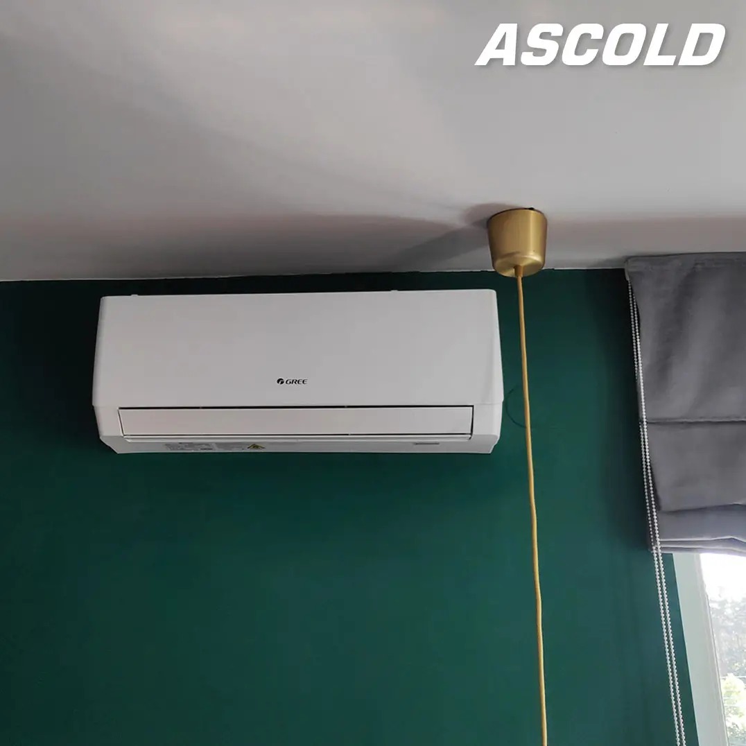 Biały klimatyzator Gree na zielonej ścianie, obok złota lampa wisząca. Widoczne okno z szarą roletą rzymską. Ascold.
