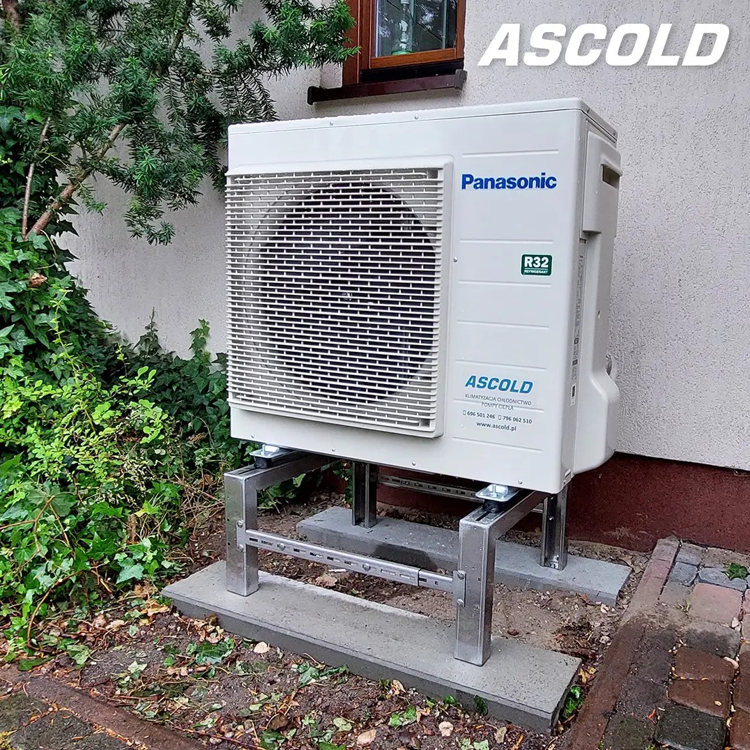 Biała pompa ciepła Panasonic na metalowej konstrukcji, zamontowana na zewnątrz budynku, otoczona zielenią. Widoczny znak firmowy Ascold. Ujęcie z dołu.