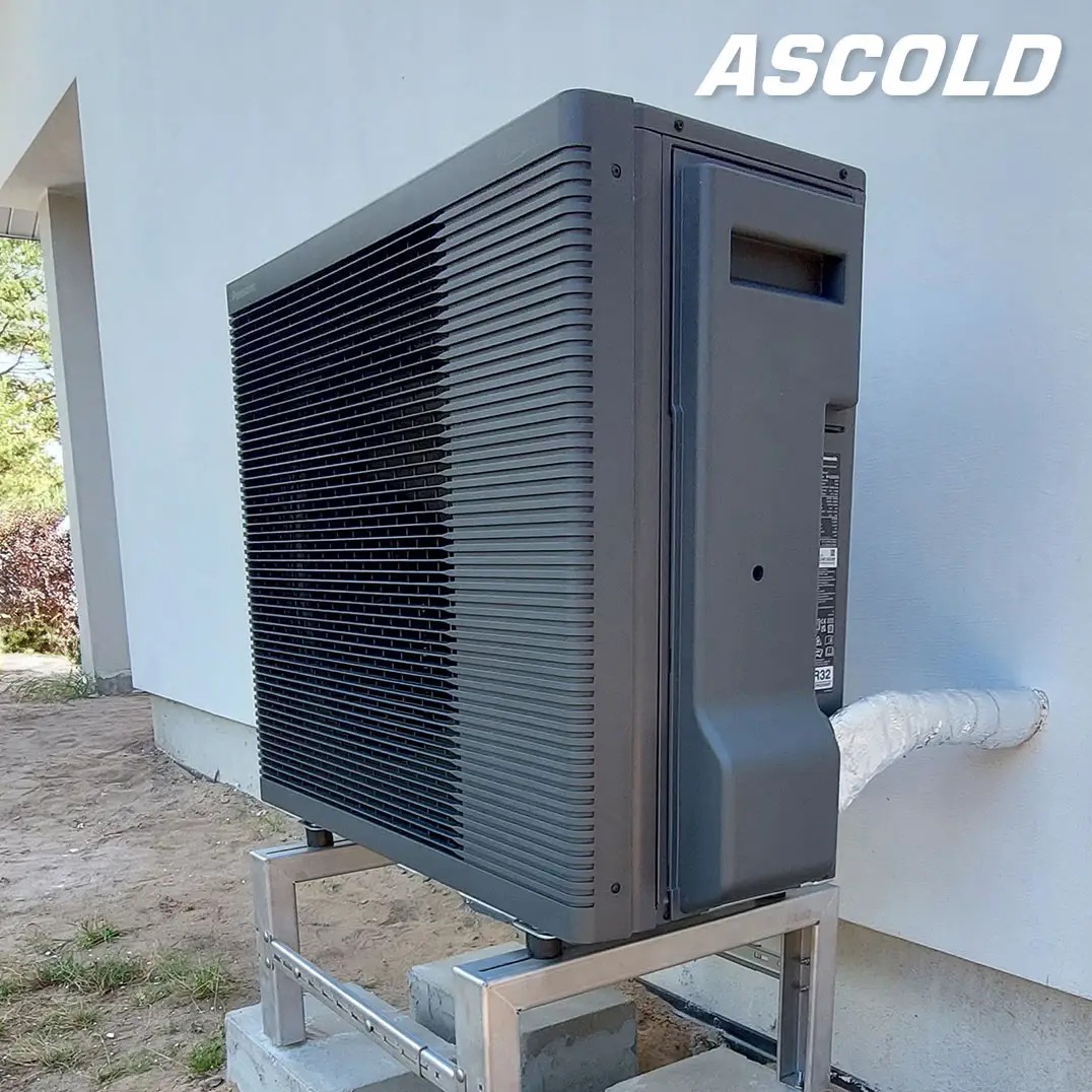 Szara pompa ciepła Ascold zamontowana na metalowej konstrukcji przy białej ścianie budynku. Widoczne przyłącza i fragment trawnika.