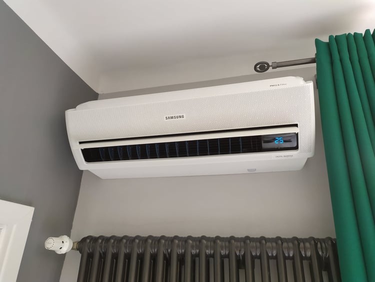 Biały klimatyzator Samsung zamontowany na szarej ścianie nad ciemnym, żeliwnym grzejnikiem, obok zielonych zasłon.