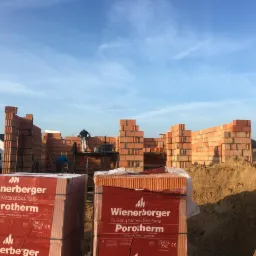 Wznoszone mury domu z czerwonej cegły Porotherm, widoczne palety z zapakowanymi pustakami ceramicznymi Wienerberger, dwóch pracowników na rusztowaniu, błękitne niebo w tle.