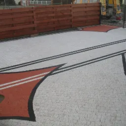 Ułożona kostka brukowa w kolorze szarym i czerwonym z czarnymi obramowaniami, tworząca wzór na podjeździe, widoczny drewniany płot i metalowa brama w tle.